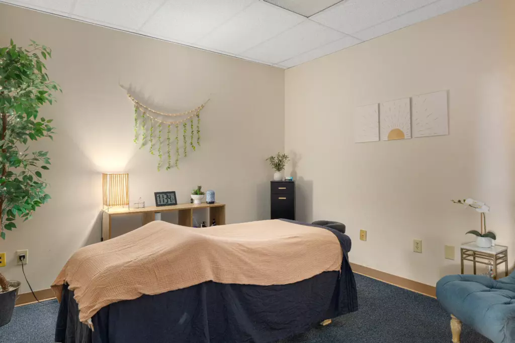 Massage Therapy – A massage therapy room with a covered massage table, soft lighting, potted plants, wall decor, and a cabinet, creating a calm and relaxing atmosphere. | Body Heal Therapy
